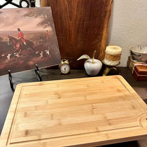 May include: A rectangular bamboo cutting board with a juice groove. The board is light brown with visible wood grain. A painting of a fox hunt, a small clock, and a decorative apple are in the background.