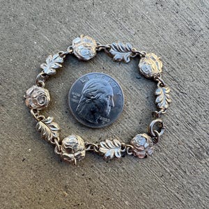 May include: A silver-toned bracelet featuring alternating rose and leaf designs. The bracelet is circular and has a clasp closure. A U.S. quarter coin is next to the bracelet for size comparison. The bracelet is on a gray surface.