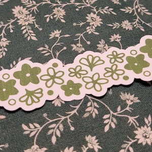 May include: Three green floral patterned stickers on a green floral fabric background. The stickers are in the shape of a banner with a scalloped edge. The stickers have a white outline and green flowers.