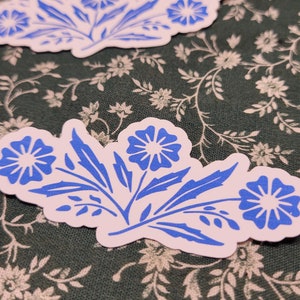 May include: Two blue and white floral stickers with a simple, stylized design. The stickers feature blue flowers with white centers and white stems and leaves.