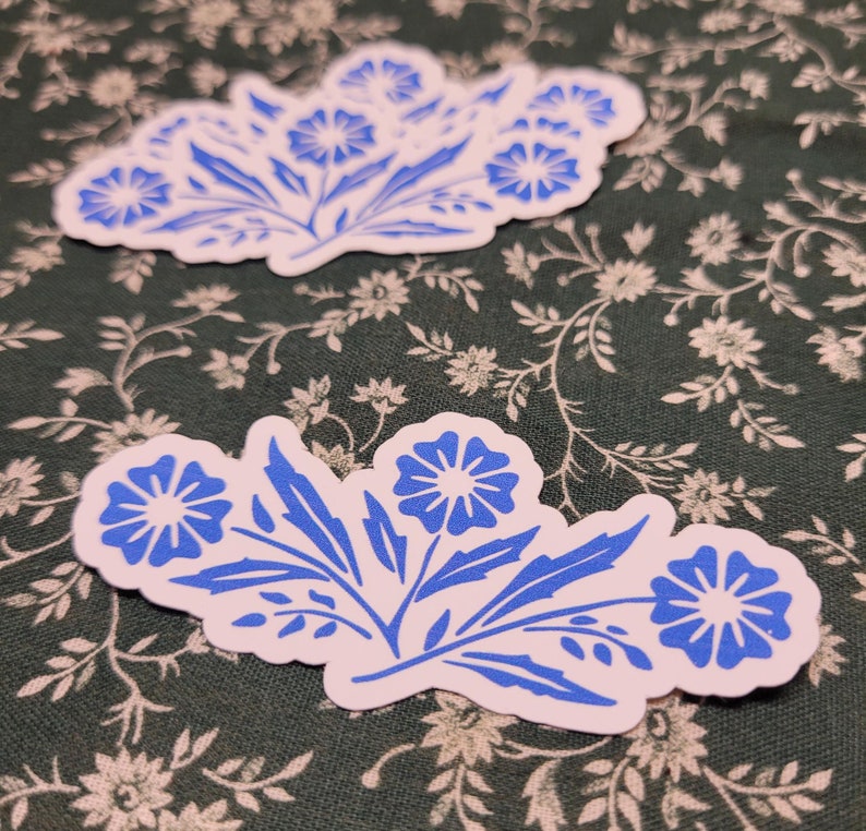 Pu&ograve; includere: Due adesivi floreali blu e bianchi con un design semplice e stilizzato. Gli adesivi presentano fiori blu con centri bianchi e steli e foglie bianche.