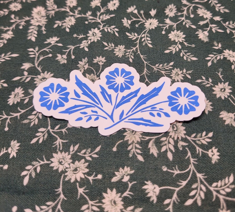 Pu&ograve; includere: Un adesivo bianco con un motivo floreale blu. L'adesivo presenta tre fiori stilizzati con cinque petali ciascuno e steli con foglie.