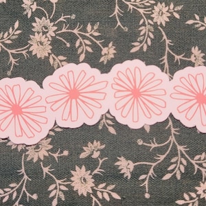 May include: A row of six pink paper flower stickers with a white outline. The stickers are arranged in a line on a dark green fabric with a floral pattern.