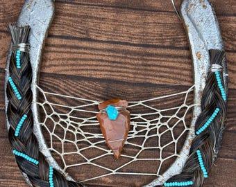 Puede incluir: Un atrapasueños de herradura plateada con detalles de crin trenzado y cuentas turquesas. Una punta de flecha marrón está centrada dentro de una telaraña blanca, con una piedra turquesa adjunta.