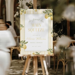 May include: A welcome sign for a bridal shower, displayed on a wooden easel. The sign has a yellow and white striped design with lemon and leaf illustrations. Text reads "She found her main squeeze" and welcomes guests to Olivia Wilson's bridal shower.