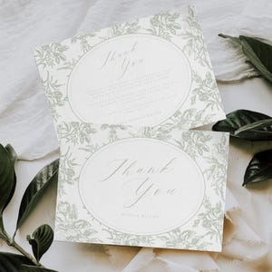 May include: Two white thank you cards with a floral design in a soft green hue. The cards feature an oval frame with the words "Thank You" in elegant script. Additional text is visible within the oval, along with names.