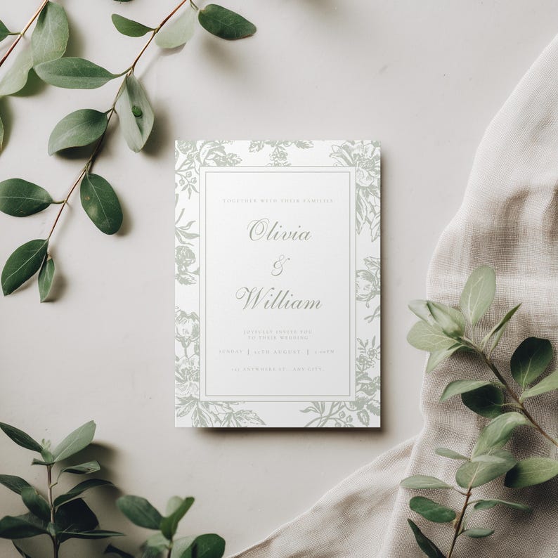 May include: A rectangular wedding invitation with floral accents and the names "Olivia & William" in elegant script. The invitation is white with a light green floral border and text. Greenery and a neutral fabric surround the invitation.