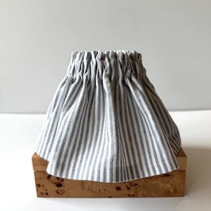 May include: A gathered fabric lampshade in a blue and white striped pattern. The shade is made of a lightweight material and has a flared shape. The lampshade is sitting on a wooden box.