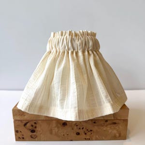 May include: Cream-colored lampshade with vertical ribbed texture, gathered at the top. The lampshade sits atop a wooden box with a burl wood grain pattern. The background is a plain white wall.