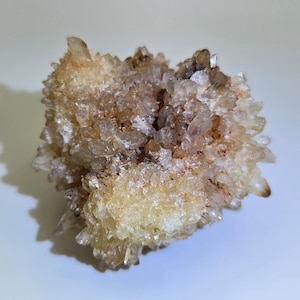 May include: A cluster of quartz crystals with a light brown and yellow hue. The crystals are clustered together, creating a textured, spiky appearance. The quartz formation is on a white surface.