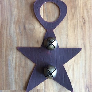 May include: A purple leather star-shaped door knocker with two brass bells attached. The knocker is shaped like a star with a rounded top and a hole in the center. The bells are round and have a cross-shaped design.