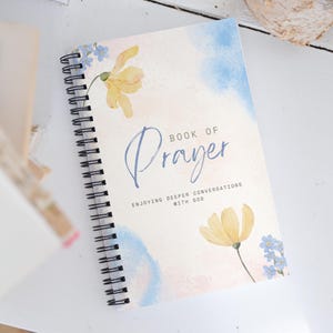 May include: A spiral bound journal with a watercolor background of blue and pink. The cover features yellow flowers and the text "Book of Prayer" and "Enjoying Deeper Conversations with God".