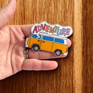 May include: A colorful sticker featuring a yellow and white vintage van with blue windows. The word "ADVENTURE" is written above the van in rainbow colors. The sticker is held in a hand against a wooden background.