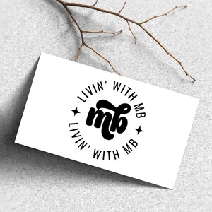 May include: A white card with the text "LIVIN' WITH MB" in a circular design. The center features the letters "mb" in a bold, cursive font. Small star shapes are on either side of the text. A twig is in the background.