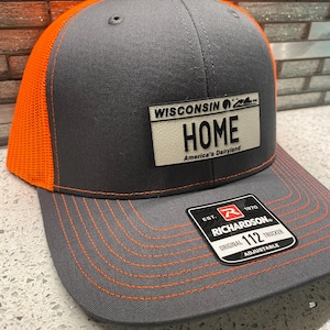May include: A gray and orange trucker hat with the text "WISCONSIN HOME America's Dairyland" on the front. The hat has an orange mesh back and brim, with orange stitching. A black and white tag is attached.