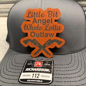 May include: A gray trucker hat with a brown leather patch that says "Little Bit Angel Whole Lotta Outlaw" with two crossed pistols. The hat has a black mesh back and a Richardson 112 adjustable strap.