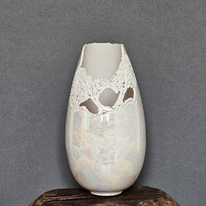 May include: A white ceramic vase with a pearlescent glaze and a cutout design of trees. The vase is shaped like an egg and has a wide opening at the top.