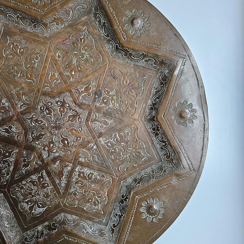 May include: A round, metallic, bronze-toned tray featuring an intricate, engraved eight-pointed star pattern. The surface is decorated with detailed floral motifs and small, circular flower-like stamps along the outer rim.