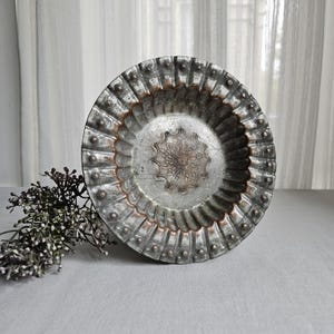 May include: A round, metallic bowl with a silver and copper-toned finish. The bowl features a fluted, scalloped rim with small raised dots and a central medallion with an intricate, embossed geometric pattern. The surface shows a weathered texture.