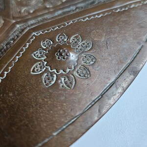 May include: A copper tray featuring an embossed floral motif at its center, surrounded by intricate geometric patterns and scalloped borders. The metal surface displays a weathered patina with visible oxidation and fine etched detailing.