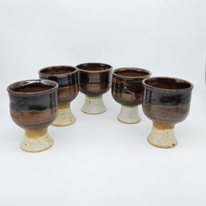 May include: A set of five handmade ceramic goblets. Each goblet features a dark brown interior, a band of brown, and a speckled beige base. The goblets have a unique, rustic appearance, perfect for serving beverages.