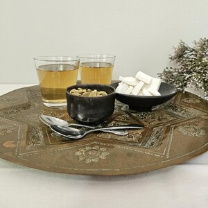 May include: A large, round, engraved brass tray holds two clear glass tumblers filled with amber liquid, a small black bowl containing green cardamom pods, a black dish with white sugar cubes, and two silver-toned metal spoons.