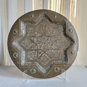 May include: A round brass tray featuring an intricate eight-pointed star design at the center, surrounded by detailed floral engravings and small circular flower motifs along the outer rim, all finished with a weathered metallic patina.