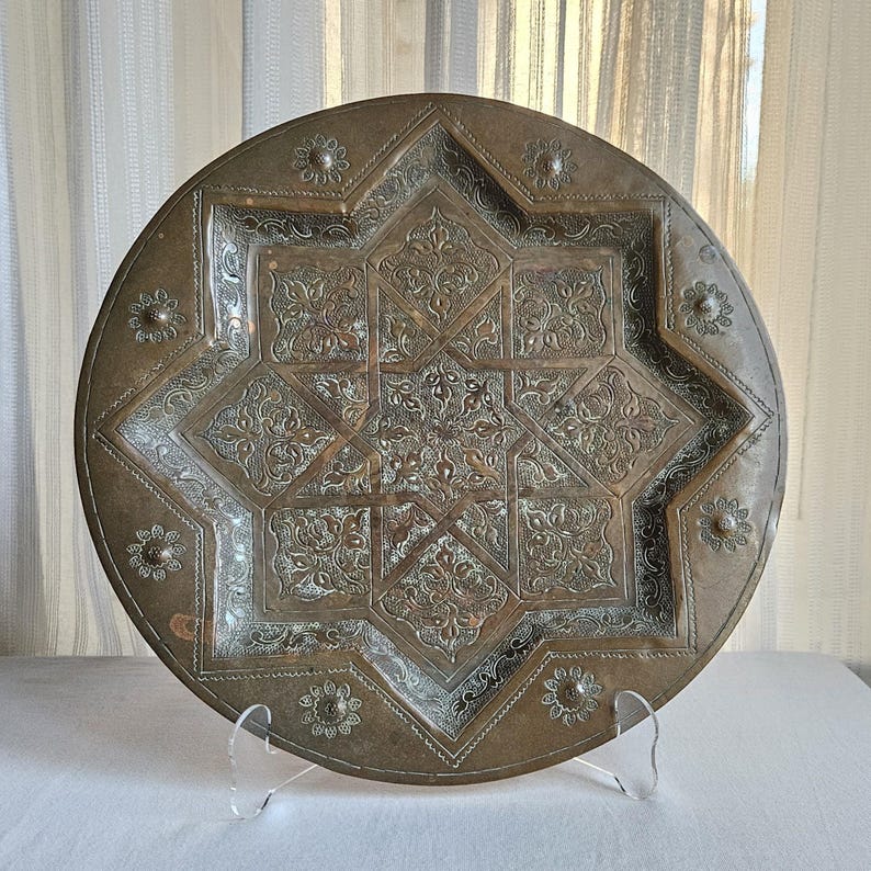 May include: A round brass tray featuring an intricate eight-pointed star design at the center, surrounded by detailed floral engravings and small circular flower motifs along the outer rim, all finished with a weathered metallic patina.