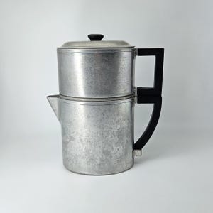 May include: A two-tiered aluminum coffee pot with a brushed metal finish. The cylindrical vessel features a small spout on the lower section, a flat lid with a black knob, and two stacked black rectangular handles attached to the side.