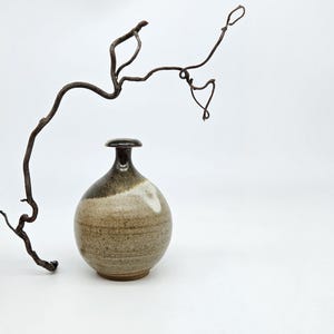 May include: A small, round ceramic vase with a narrow neck and a flat rim. The vase features a speckled, light tan base that transitions into a dark brown glaze at the top. A thin, curved, dark brown twig arches gracefully over the vase.
