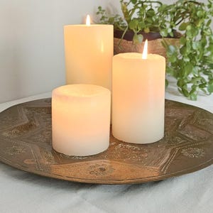 May include: Three white cylindrical pillar candles with lit flames are arranged on a large, round, antique-style brass tray featuring an intricate, embossed star pattern and floral details across its metallic surface.