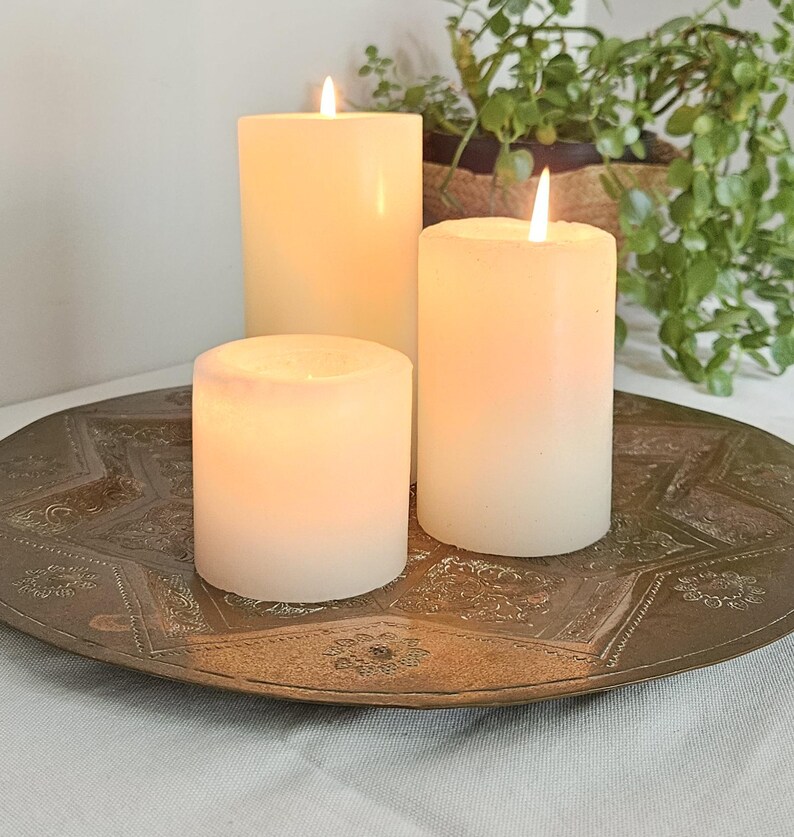 May include: Three white cylindrical pillar candles with lit flames are arranged on a large, round, antique-style brass tray featuring an intricate, embossed star pattern and floral details across its metallic surface.