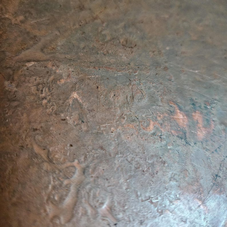 May include: A close-up view of a weathered copper surface featuring a dull, oxidized metallic finish with visible scratches, small indentations, and irregular patches of exposed reddish-brown copper showing through the aged, dark patina.