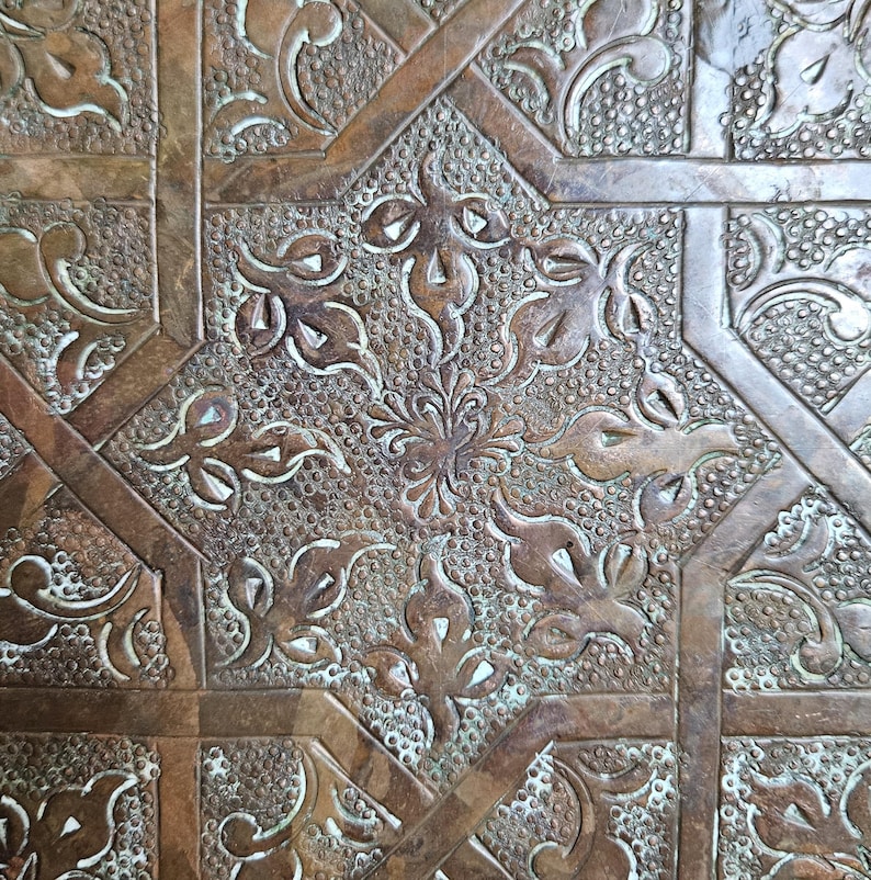 May include: A brown wooden surface featuring an intricate carved geometric pattern. The design consists of repeating star shapes and floral motifs, with a textured background of small raised dots and smooth, recessed lines throughout.