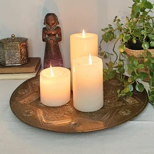 May include: Three white pillar candles with glowing flames are arranged on a round, etched brass tray. The tray features intricate floral and geometric patterns. A small wooden figurine and a metallic box sit nearby.