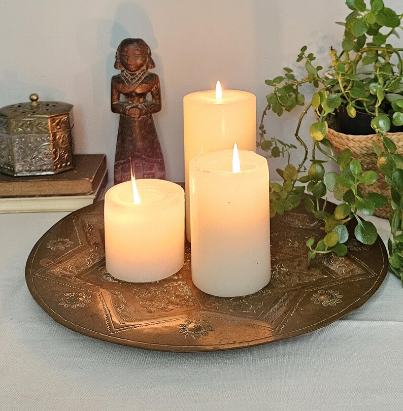 May include: Three white pillar candles with glowing flames are arranged on a round, etched brass tray. The tray features intricate floral and geometric patterns. A small wooden figurine and a metallic box sit nearby.