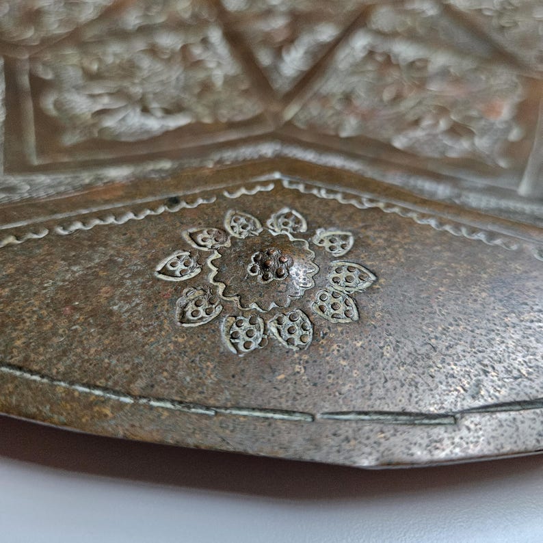May include: A metallic tray with a weathered bronze-toned finish featuring intricate hand-engraved floral patterns and geometric borders. The surface displays a central flower motif surrounded by detailed etched lines and textured metalwork.