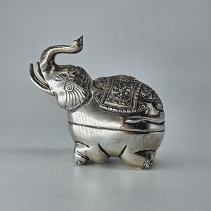 May include: A silver-colored metal elephant figurine with a raised trunk and detailed embossed patterns on its back and ears. The elephant has a horizontal seam across its midsection, indicating it is a lidded container or box.
