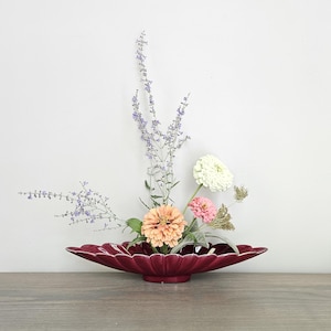 May include: A floral arrangement featuring a deep red, scalloped-edge ceramic bowl filled with colorful flowers and delicate sprigs of lavender. The flowers include peach, pink, and white zinnias, creating a vibrant and natural display. The arrangement sits on a wooden surface.