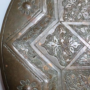 May include: A circular bronze-toned metal tray featuring intricate embossed floral and geometric patterns. The surface displays a weathered patina with textured stippled backgrounds and raised, smooth-edged botanical motifs.