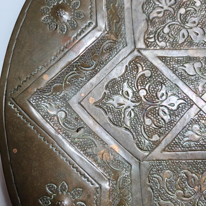 May include: A circular bronze-toned metal tray featuring intricate embossed floral and geometric patterns. The surface displays a weathered patina with textured stippled backgrounds and raised, smooth-edged botanical motifs.
