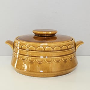 May include: A brown ceramic casserole dish with a lid. The dish has two handles and a decorative scalloped pattern around the body. The lid has a small, round knob. The dish is a warm, earthy tone.