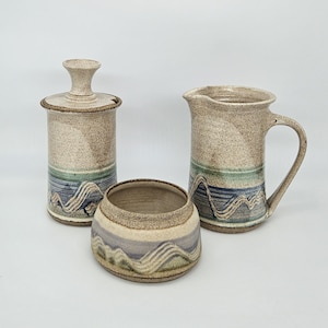 May include: A set of three ceramic kitchenware pieces in a rustic style. The pieces are a cream-colored pitcher with a handle, a cream-colored sugar bowl with a lid, and a cream-colored bowl. All three pieces have a blue, green, and brown wavy design.