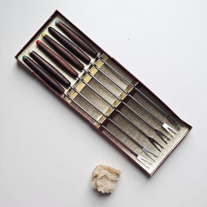 May include: A set of fondue forks in a red box. The forks have dark wooden handles with colored tips: green, red, yellow, and blue. The tines are silver. A small piece of bread sits next to the box.