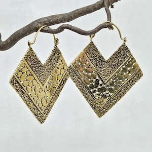 May include: A pair of brass-colored, geometric earrings with a textured surface and a V-shaped design. The earrings are diamond-shaped with a central band of small, rounded shapes. They hang from a simple hook and are displayed on a brown branch.