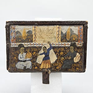 May include: A wooden box with a hinged lid, painted with a scene of three figures in a garden. The figures are wearing colorful clothing and are surrounded by flowers and foliage. The box is decorated with a geometric pattern and has a metal latch.
