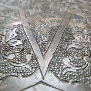May include: A metallic surface featuring an intricate, embossed pattern of raised floral motifs and geometric lines. The design includes textured, stippled backgrounds within triangular and curved sections, finished in a tarnished bronze tone.