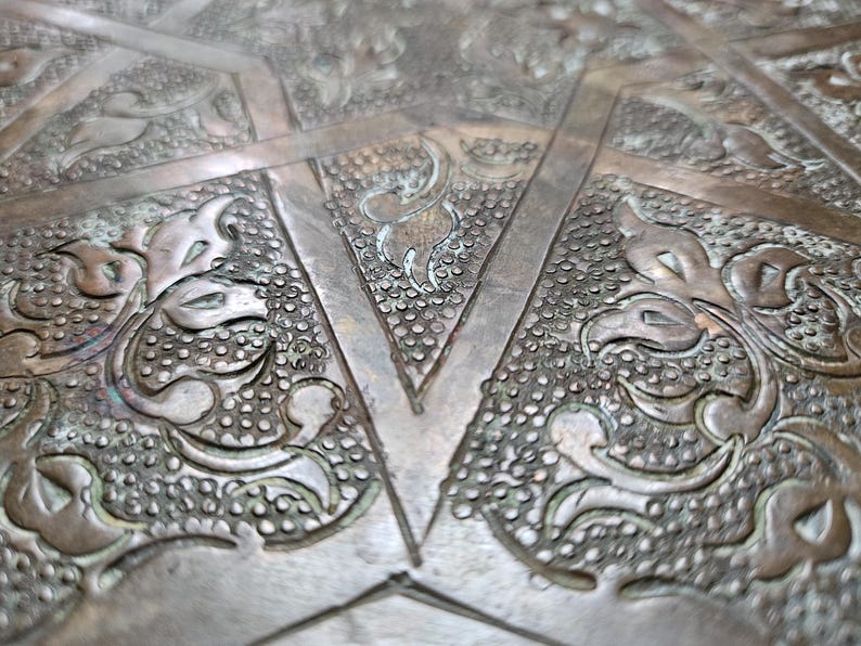 May include: A metallic surface featuring an intricate, embossed pattern of raised floral motifs and geometric lines. The design includes textured, stippled backgrounds within triangular and curved sections, finished in a tarnished bronze tone.