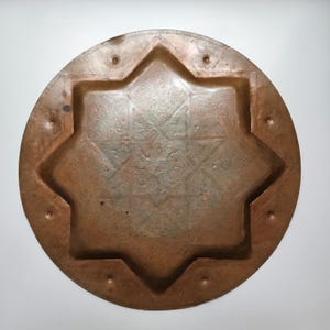 May include: A circular copper tray featuring a recessed eight-pointed star design in the center. The metal surface displays a weathered, oxidized patina with small circular indentations spaced evenly around the outer rim of the tray.