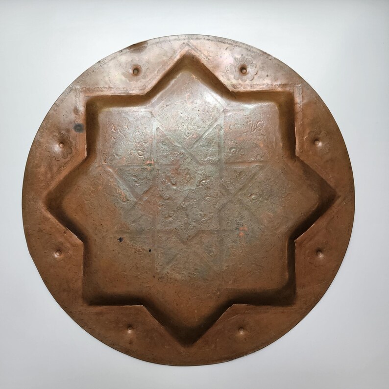May include: A circular copper tray featuring a recessed eight-pointed star design in the center. The metal surface displays a weathered, oxidized patina with small circular indentations spaced evenly around the outer rim of the tray.